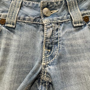 May include: Light blue denim jeans featuring prominent white contrast stitching, a silver-toned metal button at the waist with embossed text, and dark brown circular rivets at the corners of the front pockets and belt loops.