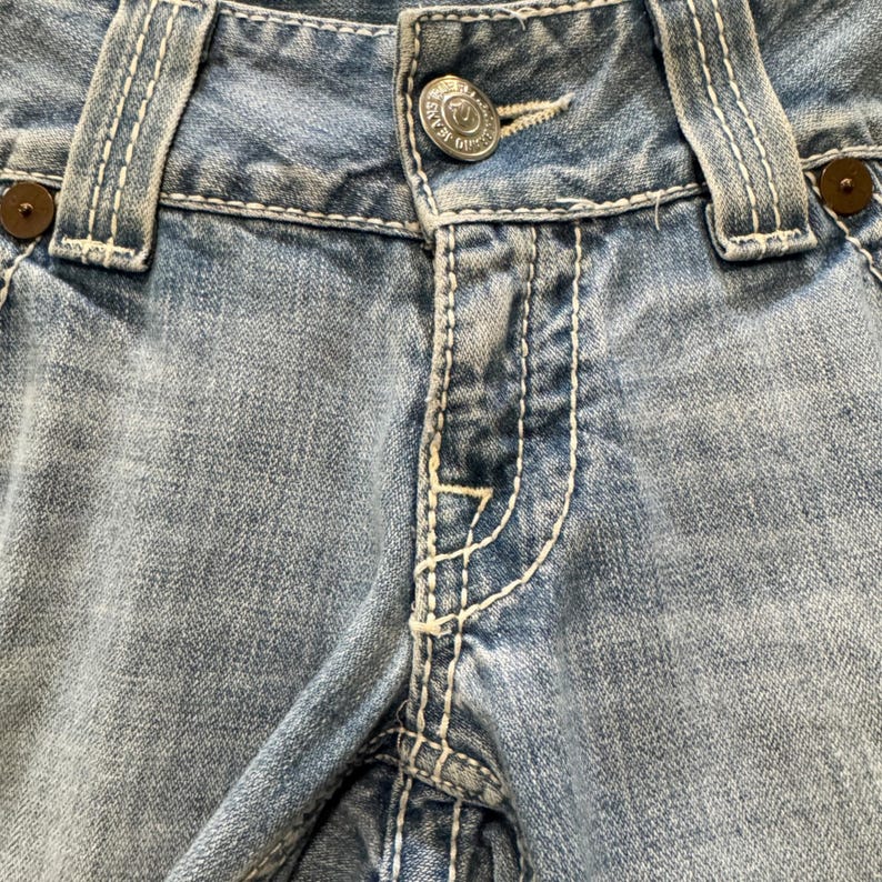 May include: Light blue denim jeans featuring prominent white contrast stitching, a silver-toned metal button at the waist with embossed text, and dark brown circular rivets at the corners of the front pockets and belt loops.