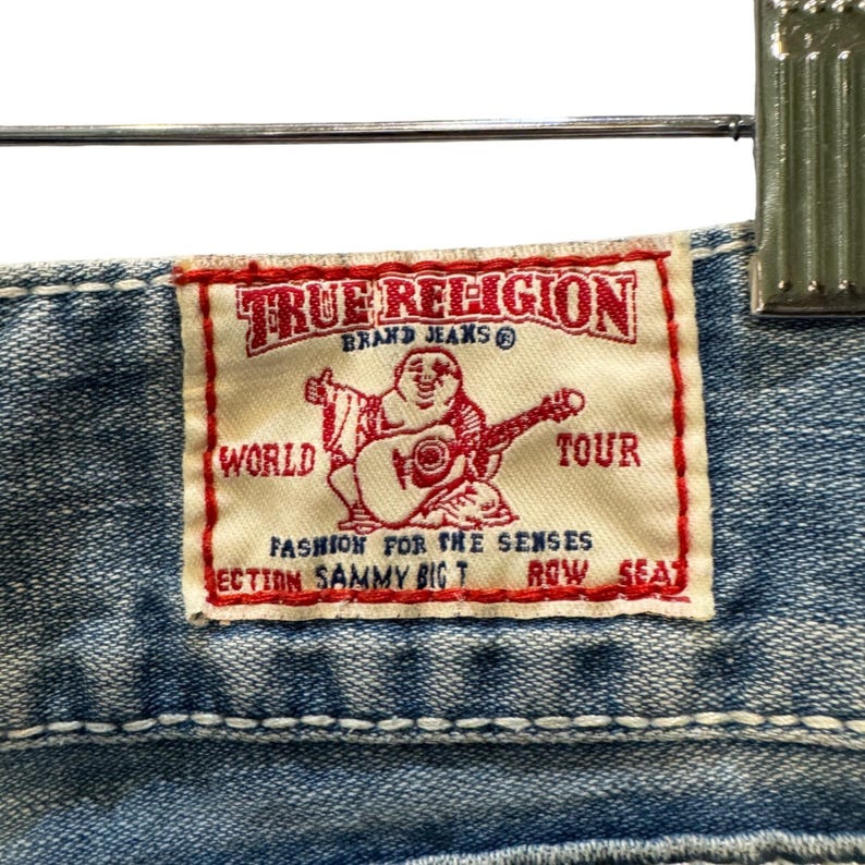 May include: A light blue denim fabric featuring a rectangular white True Religion brand label with red stitching and text. The label displays a graphic of a seated figure playing a guitar with the words True Religion Brand Jeans World Tour.