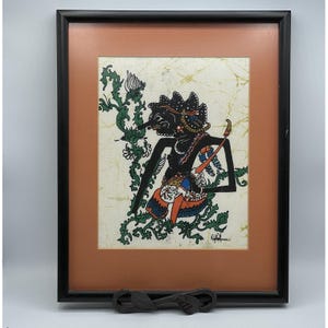 May include: A framed artwork featuring a black wayang kulit shadow puppet silhouette against a cream-colored batik background with green floral motifs. The figure wears orange and blue patterned garments. A signature is visible in the bottom right.