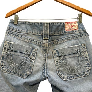 May include: Light blue denim jeans featuring thick white contrast stitching, two back pockets with a signature horseshoe design, and a red True Religion brand label on the waistband. The jeans are displayed on a wooden hanger.