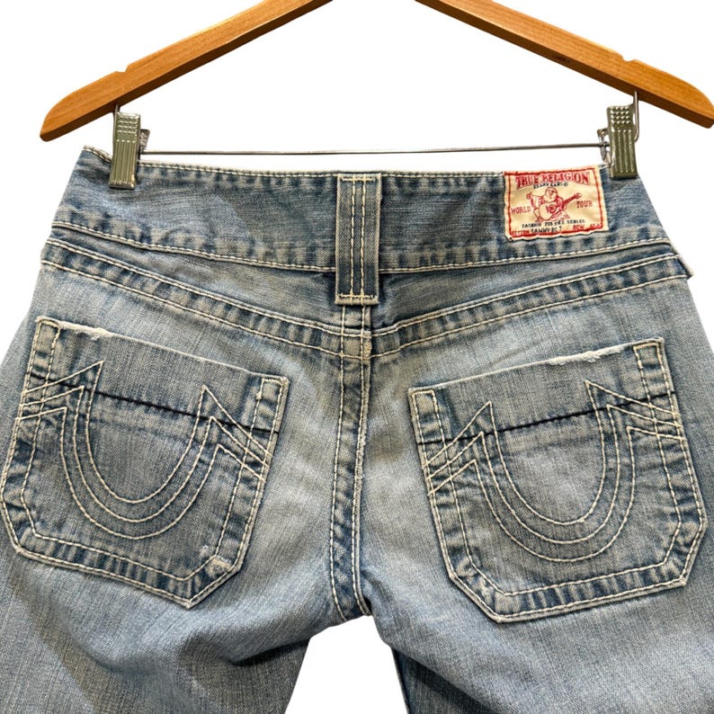 May include: Light blue denim jeans featuring thick white contrast stitching, two back pockets with a signature horseshoe design, and a red True Religion brand label on the waistband. The jeans are displayed on a wooden hanger.