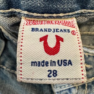 May include: A white fabric clothing tag with red and blue text and a red horseshoe logo. The tag reads TRUE RELIGION BRAND JEANS, made in USA, and the number 28. It is stitched onto blue denim fabric with thick white contrast stitching.