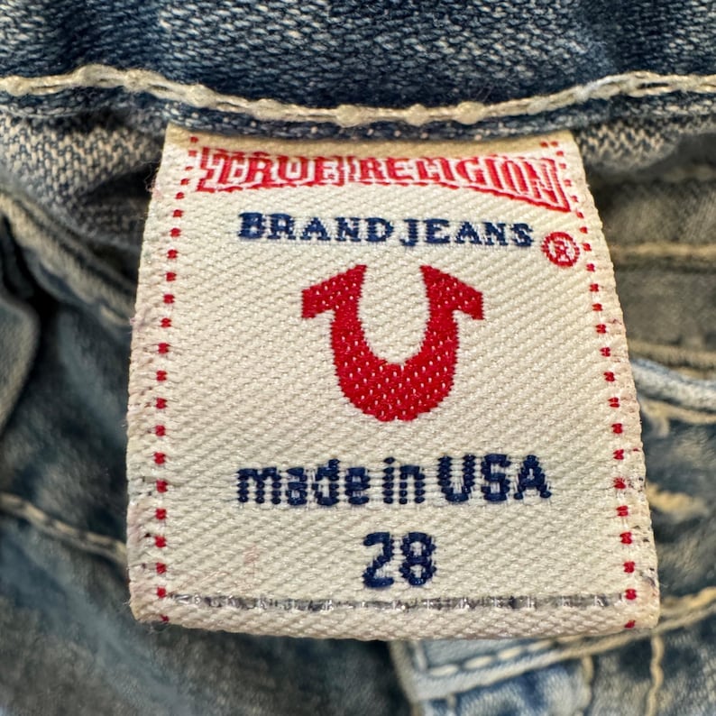 May include: A white fabric clothing tag with red and blue text and a red horseshoe logo. The tag reads TRUE RELIGION BRAND JEANS, made in USA, and the number 28. It is stitched onto blue denim fabric with thick white contrast stitching.
