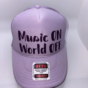 Könnte beinhalten: Eine hellviolette Trucker-Cap mit dem schwarzen Glitzertext Music ON World OFF auf der vorderen Schaumstoffplatte. Die Kappe verfügt über ein Netzrückenteil, einen gebogenen Schirm und ein weißes Etikett mit dem Markennamen OTTO und Textdetails.