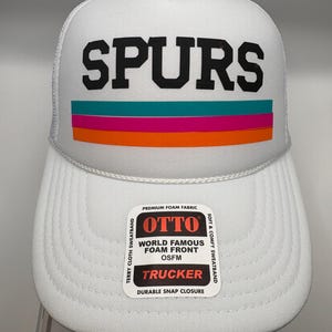 May include: A white trucker hat featuring the word SPURS in black block letters above three horizontal stripes in teal, magenta, and orange. A rectangular product label is attached to the brim with the text OTTO, WORLD FAMOUS FOAM FRONT, OSFM, TRUCKER, and other details.