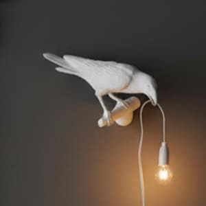 Könnte beinhalten: Eine weiße, vogelartige Wandlampe mit einer hängenden Glühbirne. Der Vogel sitzt auf einem Ast, mit der Glühbirne darunter. Die Lampe ist an einer dunkelgrauen Wand montiert, mit einem goldenen Dekorationselement oben links.