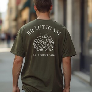 K&ouml;nnte beinhalten: Ein olivgr&uuml;nes T-Shirt mit einer wei&szlig;en Grafik auf der R&uuml;ckseite. Das Design zeigt zwei ansto&szlig;ende Bierkr&uuml;ge mit Schaum, das Wort BR&Auml;UTIGAM dar&uuml;ber gebogen und das Datum 08. AUGUST 2026 in einer serifenlosen Schrift unter den Kr&uuml;gen.