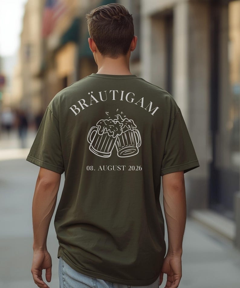 K&ouml;nnte beinhalten: Ein olivgr&uuml;nes T-Shirt mit einer wei&szlig;en Grafik auf der R&uuml;ckseite. Das Design zeigt zwei ansto&szlig;ende Bierkr&uuml;ge mit Schaum, das Wort BR&Auml;UTIGAM dar&uuml;ber gebogen und das Datum 08. AUGUST 2026 in einer serifenlosen Schrift unter den Kr&uuml;gen.