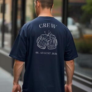 K&ouml;nnte beinhalten: Ein dunkelblaues T-Shirt mit einer wei&szlig;en Strichzeichnung von zwei ansto&szlig;enden Bierkr&uuml;gen auf der R&uuml;ckseite. &Uuml;ber den Kr&uuml;gen steht der Text CREW und darunter der Text 08. AUGUST 2026. Das T-Shirt hat kurze &Auml;rmel und einen Rundhalsausschnitt.
