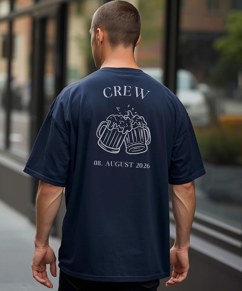 K&ouml;nnte beinhalten: Ein dunkelblaues T-Shirt mit einer wei&szlig;en Strichzeichnung von zwei ansto&szlig;enden Bierkr&uuml;gen auf der R&uuml;ckseite. &Uuml;ber den Kr&uuml;gen steht der Text CREW und darunter der Text 08. AUGUST 2026. Das T-Shirt hat kurze &Auml;rmel und einen Rundhalsausschnitt.