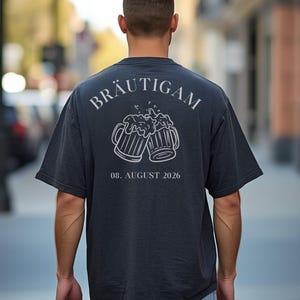 K&ouml;nnte beinhalten: Ein dunkelgraues T-Shirt mit kurzen &Auml;rmeln und einer wei&szlig;en Strichgrafik auf der R&uuml;ckseite. Das Motiv zeigt zwei ansto&szlig;ende Bierkr&uuml;ge mit Schaum, dar&uuml;ber der Text BR&Auml;UTIGAM und darunter das Datum 08. AUGUST 2026 in wei&szlig;er Schrift.