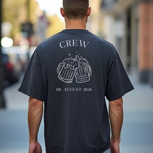 K&ouml;nnte beinhalten: Ein dunkel anthrazitfarbenes Kurzarm-T-Shirt mit einer wei&szlig;en Strichgrafik auf der R&uuml;ckseite. Das Design zeigt das Wort CREW &uuml;ber zwei ansto&szlig;enden Bierkr&uuml;gen mit Schaum sowie den Text 08. AUGUST 2026 in einer Serifenschrift unter den Kr&uuml;gen.