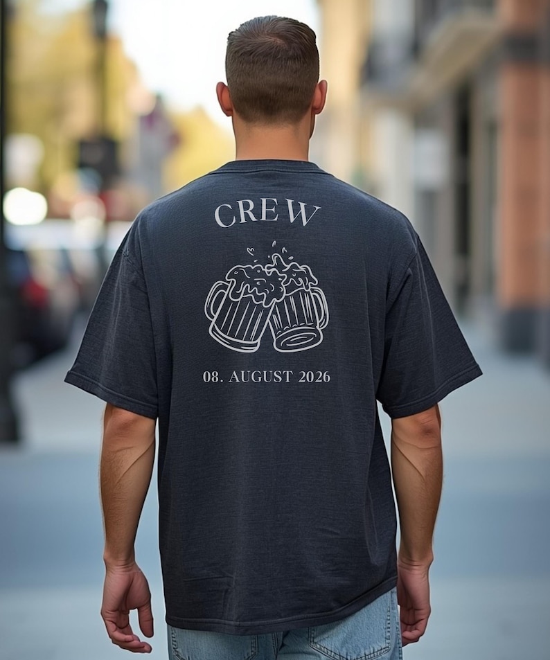 K&ouml;nnte beinhalten: Ein dunkel anthrazitfarbenes Kurzarm-T-Shirt mit einer wei&szlig;en Strichgrafik auf der R&uuml;ckseite. Das Design zeigt das Wort CREW &uuml;ber zwei ansto&szlig;enden Bierkr&uuml;gen mit Schaum sowie den Text 08. AUGUST 2026 in einer Serifenschrift unter den Kr&uuml;gen.