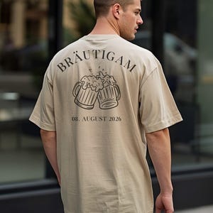 K&ouml;nnte beinhalten: Ein beiges T-Shirt mit einem schwarzen Aufdruck auf der R&uuml;ckseite. Das Motiv zeigt zwei ansto&szlig;ende Bierkr&uuml;ge mit Schaum, dar&uuml;ber der bogenf&ouml;rmige Schriftzug "BR&Auml;UTIGAM" und darunter das Datum "08. AUGUST 2026" in schwarzer Schrift.