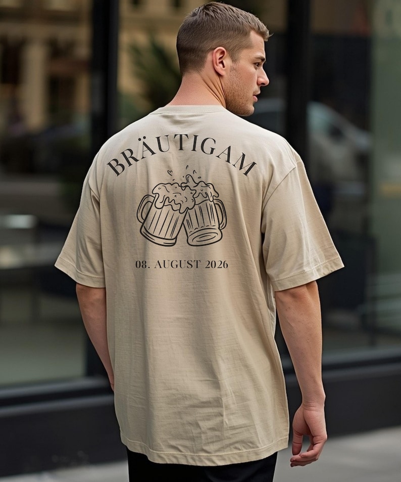 K&ouml;nnte beinhalten: Ein beiges T-Shirt mit einem schwarzen Aufdruck auf der R&uuml;ckseite. Das Motiv zeigt zwei ansto&szlig;ende Bierkr&uuml;ge mit Schaum, dar&uuml;ber der bogenf&ouml;rmige Schriftzug "BR&Auml;UTIGAM" und darunter das Datum "08. AUGUST 2026" in schwarzer Schrift.