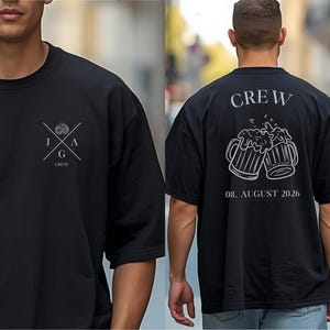 K&ouml;nnte beinhalten: Ein schwarzes T-Shirt mit einer wei&szlig;en Grafik auf der Vorderseite, die die Buchstaben J, A, G und das Wort CREW zeigt. Die R&uuml;ckseite zeigt eine gro&szlig;e wei&szlig;e Grafik von zwei ansto&szlig;enden Bierkr&uuml;gen, den Text CREW und das Datum 08. AUGUST 2026.