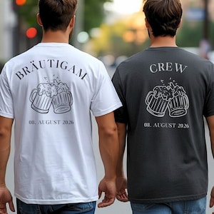 K&ouml;nnte beinhalten: Ein wei&szlig;es T-Shirt mit dem Text BR&Auml;UTIGAM und einer dunklen Grafik von zwei ansto&szlig;enden Bierkr&uuml;gen &uuml;ber dem Datum 08. AUGUST 2026, sowie ein dunkelgraues T-Shirt mit dem Text CREW und einer wei&szlig;en Grafik von zwei Bierkr&uuml;gen &uuml;ber 08. AUGUST 2026.