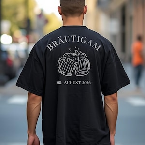 K&ouml;nnte beinhalten: Ein schwarzes Kurzarm-T-Shirt mit einer wei&szlig;en Strichzeichnung von zwei ansto&szlig;enden Bierkr&uuml;gen auf der R&uuml;ckseite. &Uuml;ber den Kr&uuml;gen steht der Text BR&Auml;UTIGAM in Serifenschrift, darunter das Datum 08. AUGUST 2026 in passender wei&szlig;er Schrift.