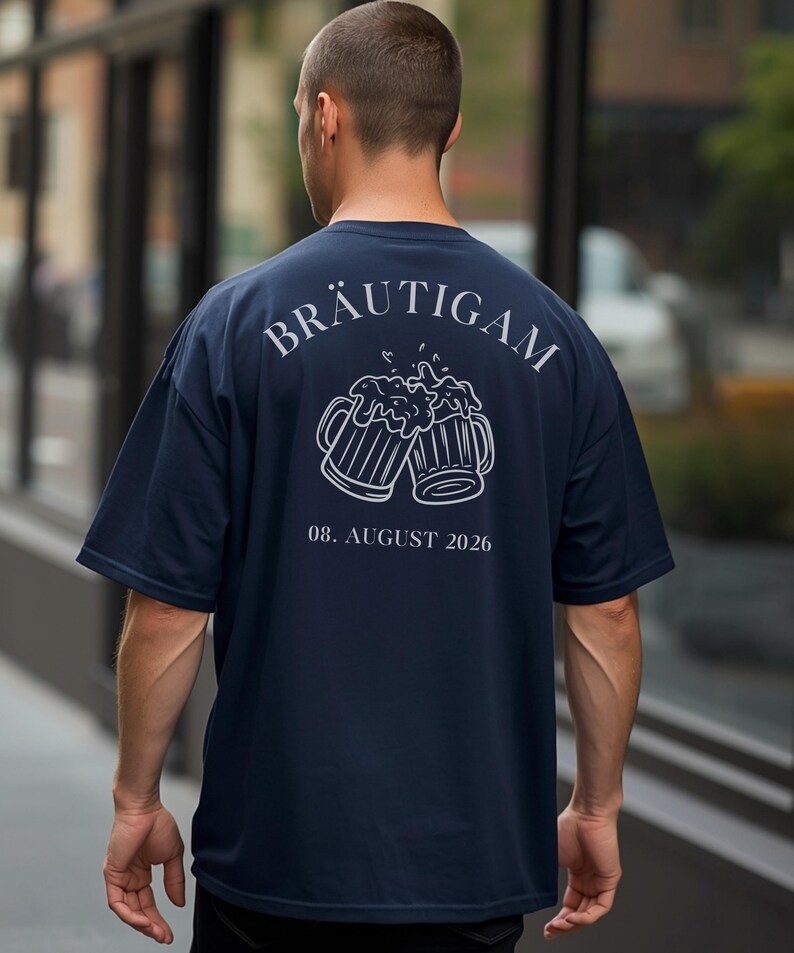 K&ouml;nnte beinhalten: Ein dunkelblaues Kurzarm-T-Shirt mit einer wei&szlig;en Linienzeichnung von zwei ansto&szlig;enden Bierkr&uuml;gen auf der R&uuml;ckseite. &Uuml;ber den Kr&uuml;gen steht der Text BR&Auml;UTIGAM in gebogener Schrift, darunter befindet sich das Datum 08. AUGUST 2026 in wei&szlig;er Schrift.