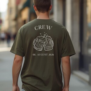 K&ouml;nnte beinhalten: Ein olivgr&uuml;nes T-Shirt mit einer wei&szlig;en Strichzeichnung von zwei ansto&szlig;enden Bierkr&uuml;gen auf der R&uuml;ckseite. Der Schriftzug "CREW" steht &uuml;ber den Kr&uuml;gen und das Datum "08. AUGUST 2026" ist darunter in einer klaren, wei&szlig;en Schriftart gedruckt.