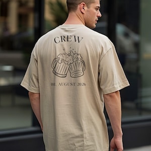 K&ouml;nnte beinhalten: Ein beiges T-Shirt mit kurzen &Auml;rmeln und einem schwarzen Aufdruck auf der R&uuml;ckseite. Das Motiv zeigt zwei ansto&szlig;ende Bierkr&uuml;ge mit Schaum, dar&uuml;ber das Wort "CREW" und darunter der deutlich gedruckte Text "08. AUGUST 2026".