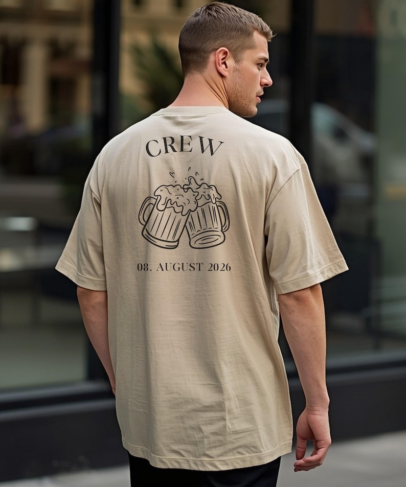 K&ouml;nnte beinhalten: Ein beiges T-Shirt mit kurzen &Auml;rmeln und einem schwarzen Aufdruck auf der R&uuml;ckseite. Das Motiv zeigt zwei ansto&szlig;ende Bierkr&uuml;ge mit Schaum, dar&uuml;ber das Wort "CREW" und darunter der deutlich gedruckte Text "08. AUGUST 2026".