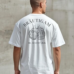 K&ouml;nnte beinhalten: Ein wei&szlig;es T-Shirt mit kurzen &Auml;rmeln und einem schwarzen Aufdruck auf der R&uuml;ckseite. Das Motiv zeigt zwei ansto&szlig;ende Bierkr&uuml;ge mit Schaum, dar&uuml;ber der in einem Bogen geschriebene Schriftzug BR&Auml;UTIGAM und darunter das Datum 08. AUGUST 2026.