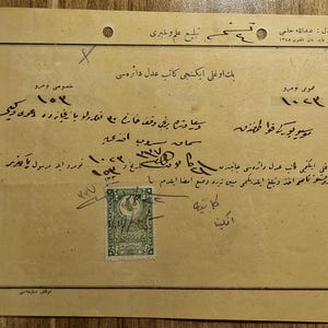 May include: A vintage off-white paper document featuring handwritten Ottoman Turkish script in black ink. The document includes printed text at the top and a rectangular green postage stamp with intricate patterns affixed to the lower section.