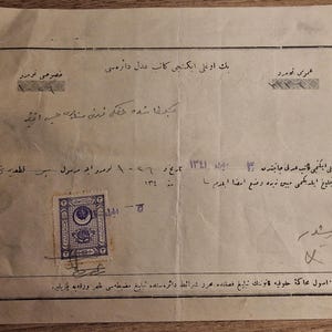 May include: A vintage document with text in a foreign language, featuring a purple stamp with an ornate design. The paper is aged, with visible creases and holes along the left edge. Handwritten notes and signatures are present, adding to its historical appearance.