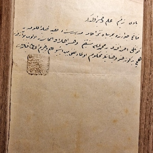 May include: A rectangular, aged, off-white paper document featuring handwritten black ink calligraphy in Ottoman Turkish script. The paper shows signs of wear, including creases and light brown staining. A square black ink stamp is visible.
