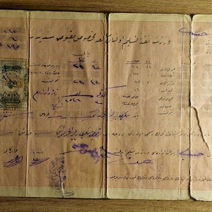 May include: A vintage document with faded text and stamps, featuring handwritten script in a foreign language. The paper is aged with visible creases and tears, suggesting historical significance. The document is displayed on a wooden surface.