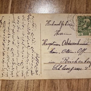 Puede incluir: Una postal vintage con texto manuscrito en varios idiomas. La postal presenta un sello postal verde y está dirigida a Hauptman Abdurrahman en Reichenberg. La tarjeta está sobre una superficie de madera.