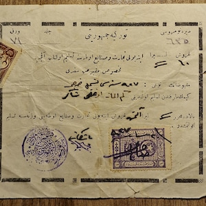 May include: A vintage off-white paper document featuring Ottoman Turkish script, black printed borders, and handwritten details. The document includes two affixed postage stamps, one brown and one purple, along with a circular blue ink stamp.
