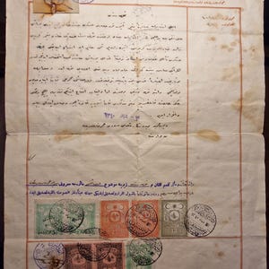 May include: An aged, off-white document featuring handwritten Arabic script, a sepia-toned portrait of a person in a suit and fez, and several colorful postage stamps with circular ink postmarks. The paper shows visible creases and brown stains.