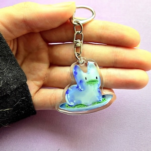 May include: A clear acrylic keychain featuring a blue bunny design with a green mouth and a light blue base. The keychain has a silver-colored metal clasp and chain. The bunny is painted with watercolor-style details.