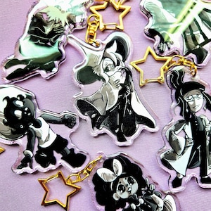 May include: A collection of anime-style character keychains in black, white, and gray. Each keychain features a unique character design with gold-colored star-shaped clasps. The keychains are displayed on a light purple surface.