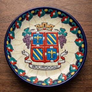 Könnte beinhalten: Dekorative Keramikplatte mit dem Wappen von Burgund. Die Platte hat eine cremefarbene Mitte und einen blauen Rand. Das Wappen enthält eine Krone, Fleur-de-Lis und das Wort "BOURGOGNE".