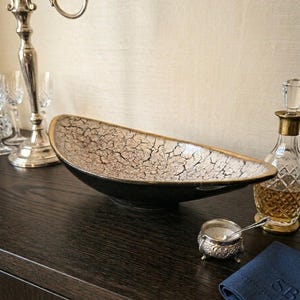 Peut inclure: Un bol décoratif noir et doré avec un intérieur craquelé et texturé. Le bol est allongé et posé sur une surface en bois sombre, à côté d'un candélabre argenté, d'une petite coupelle argentée et d'une carafe.