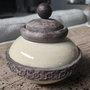 Può includere: Un barattolo in ceramica con coperchio, caratterizzato da una base color crema con una finitura smaltata a craquelé. Il barattolo presenta un bordo testurizzato marrone scuro con motivo floreale e un coperchio con pomello sferico.