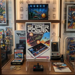 以下が含まれることがあります： ヘリコプターの戦闘シーンが描かれた、ヴィンテージのEntex Super Cobra電子アーケードゲームの箱。箱にはSuper Cobraのタイトル、ジョイスティックコントローラー、赤、白、青のストライプがデザインされており、ディスプレイ棚の上に置かれています。