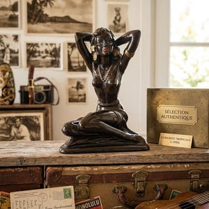 Op de afbeelding: Een donkerbruin art deco beeldje van een vrouw met haar armen achter haar hoofd, met gouden oorbellen en een ketting. Het beeldje staat op een houten oppervlak met een vintage koffer, ansichtkaarten en een ukelele. De achtergrond bevat ingelijste vintage foto's.