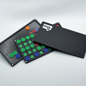 May include: A black puzzle game with colorful circular pieces and a black card with a white graphic. The game includes a black tray with various colored pieces, including green, blue, red, and orange. The card has a white design.