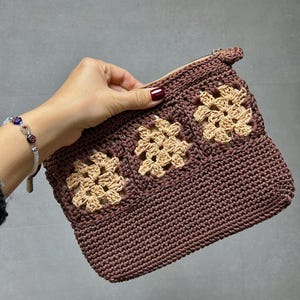 Op de afbeelding: Een bruine gehaakte clutch met drie beige bloemmotieven. De clutch heeft een ritssluiting en wordt door een hand vastgehouden. Het haakwerk is gedetailleerd en de neutrale kleuren maken het tot een veelzijdig accessoire.