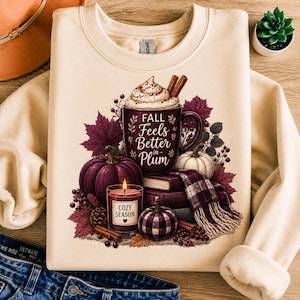 May include: A cream-colored crewneck sweatshirt featuring a graphic of a dark plum mug with whipped cream and cinnamon sticks, text reading Fall Feels Better in Plum, a candle labeled Cozy Season, pumpkins, books, and a plaid scarf.