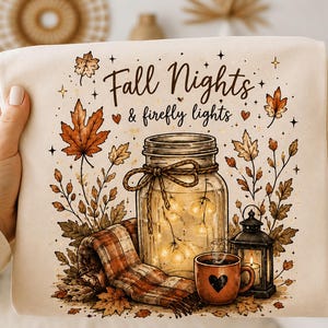 May include: A cream-colored sweatshirt featuring a graphic print with the text Fall Nights & firefly lights. The design includes a mason jar filled with glowing lights, a plaid scarf, a heart-patterned mug, a lantern, and autumn leaves.