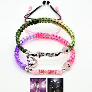 May include: Two braided cord bracelets featuring silver-toned heart charms and white rectangular beads. One bracelet is olive green with the text $AD BOYZ, the other is pink and purple with $AD GIRLZ. Both include a round bead with the number 4.