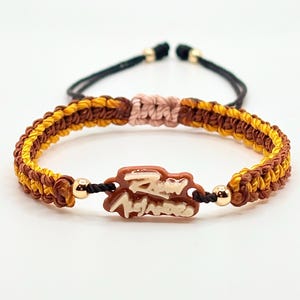Puede incluir: Una pulsera tejida con patrones de cordón alternos en amarillo mostaza y marrón oscuro. Un dije central marrón muestra texto en blanco. La pulsera incluye dos pequeñas cuentas metálicas doradas y un cierre de nudo corredizo rosa pálido.