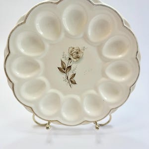 May include: A round, cream-colored ceramic egg serving plate with a scalloped edge and a metallic silver rim. The center features a detailed illustration of a single rose with brown leaves and stems, surrounded by twelve oval-shaped indentations.