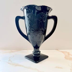 May include: Black glass vase with two handles and a square base. The vase features a heart-shaped design with embossed figures. The rim has a ruffled edge. The vase is approximately 8 inches tall.